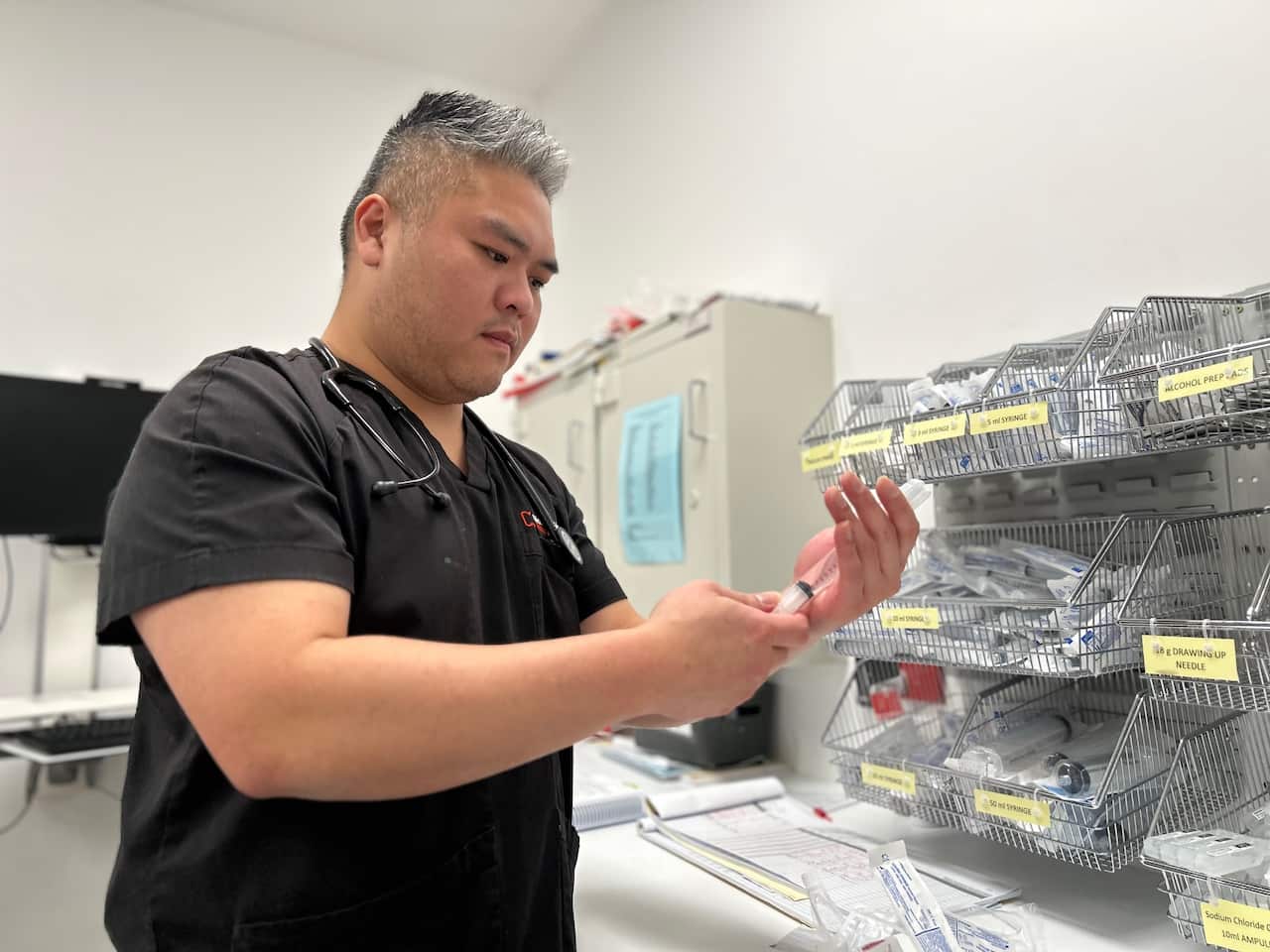A man in a black nursing uniform stands in a medical room holding a syringe.