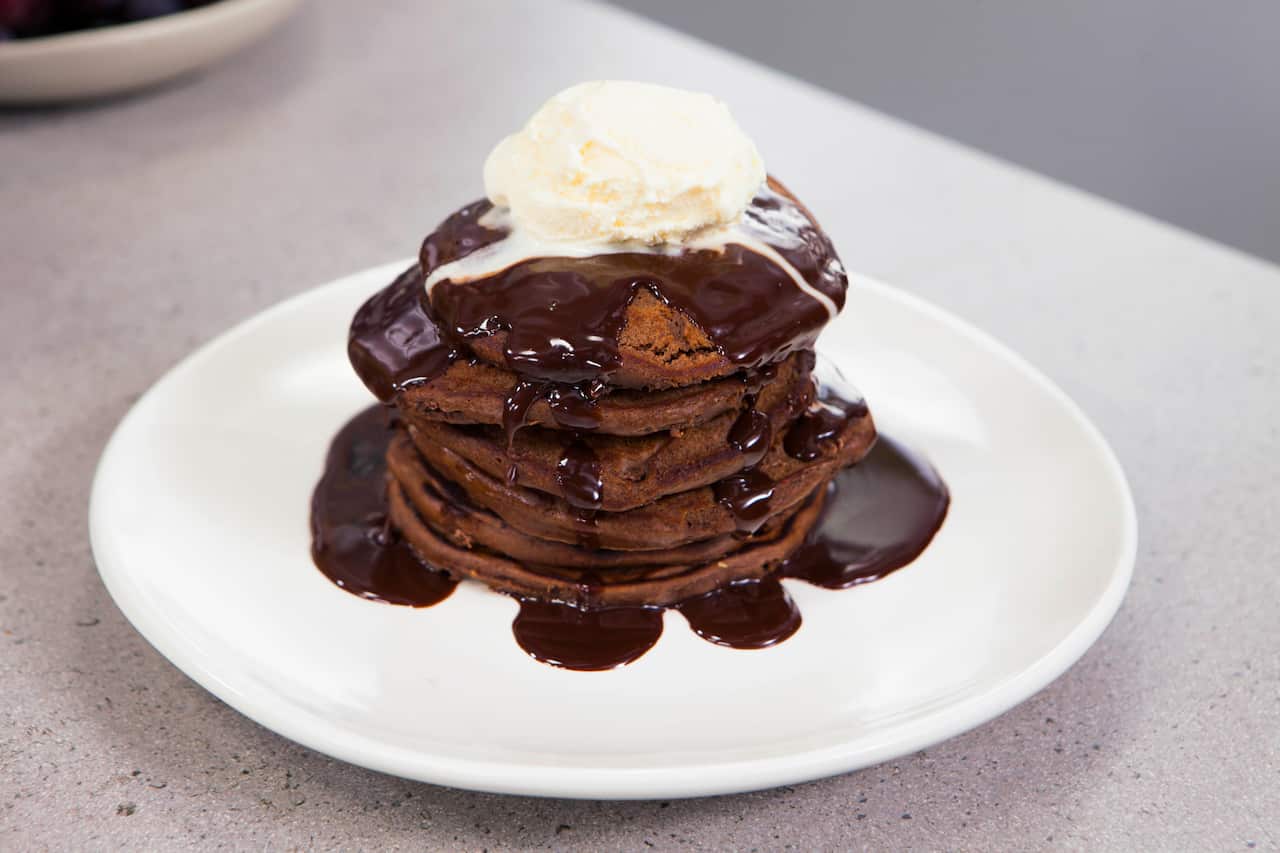 A stack of chocolate pancakes sits on a white plate, topped with ice-cream and chocolate sauce.