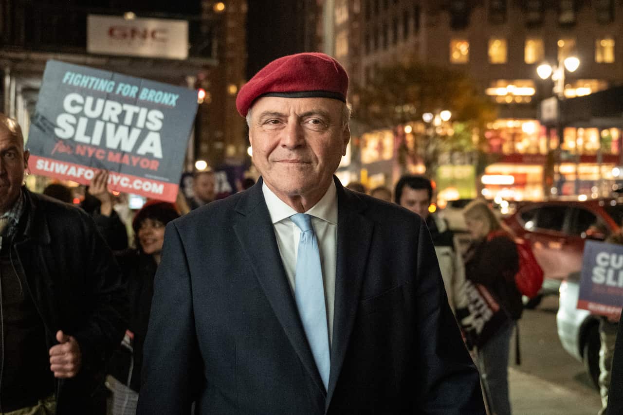 An older white man wearing a red beret and navy suit. He his standing a city street, supporters surround him holding placards saying 'Curtis Sliwa for NYC Mayor'.