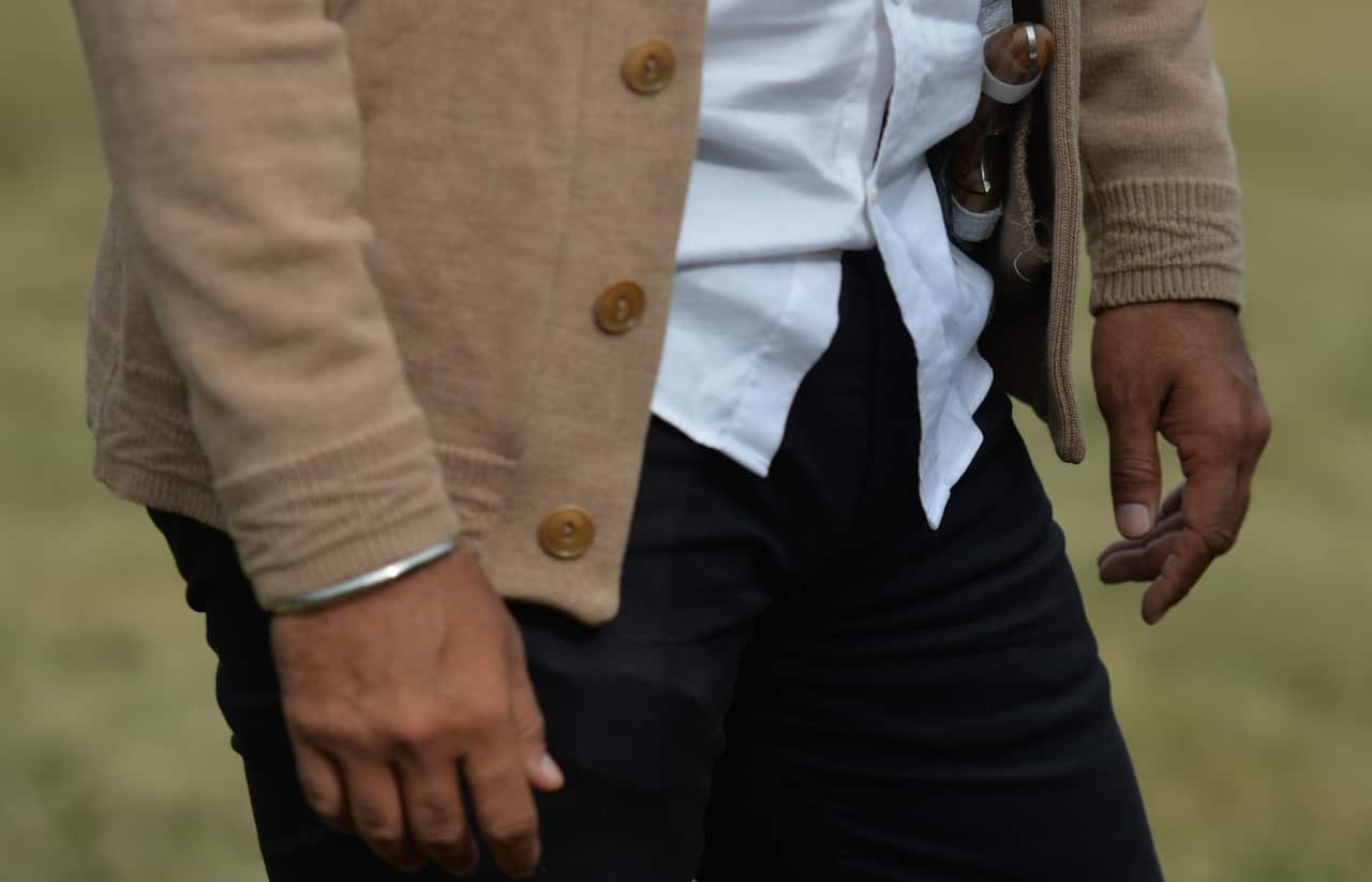 A close-up of a person wearing a tan cardigan, white shirt, and black pants, with a silver bracelet (Kara) on one wrist and the hilt of a small dagger (Kirpan) visibly tucked into their clothing on the opposite side.