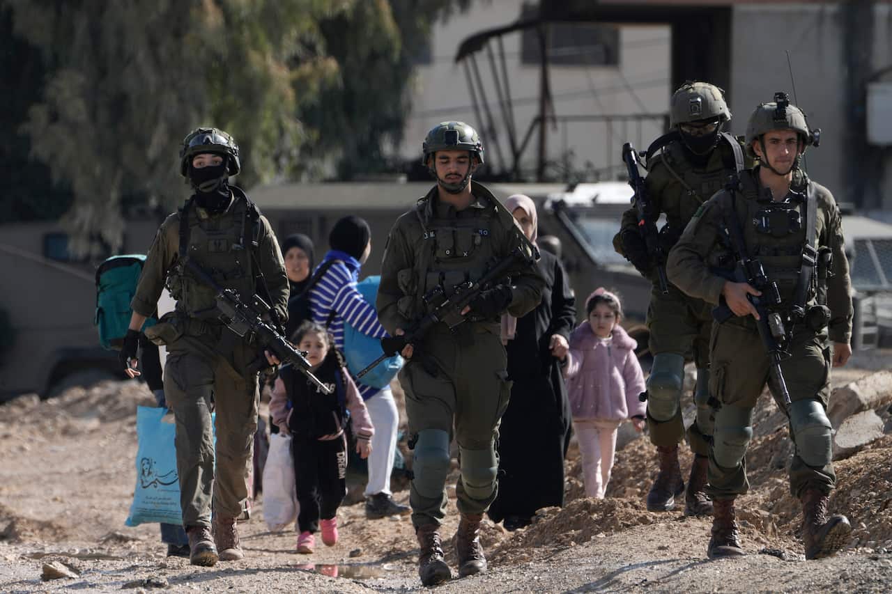 Soldiers in uniform holding guns march in front of young Palestinian children and their mothers.