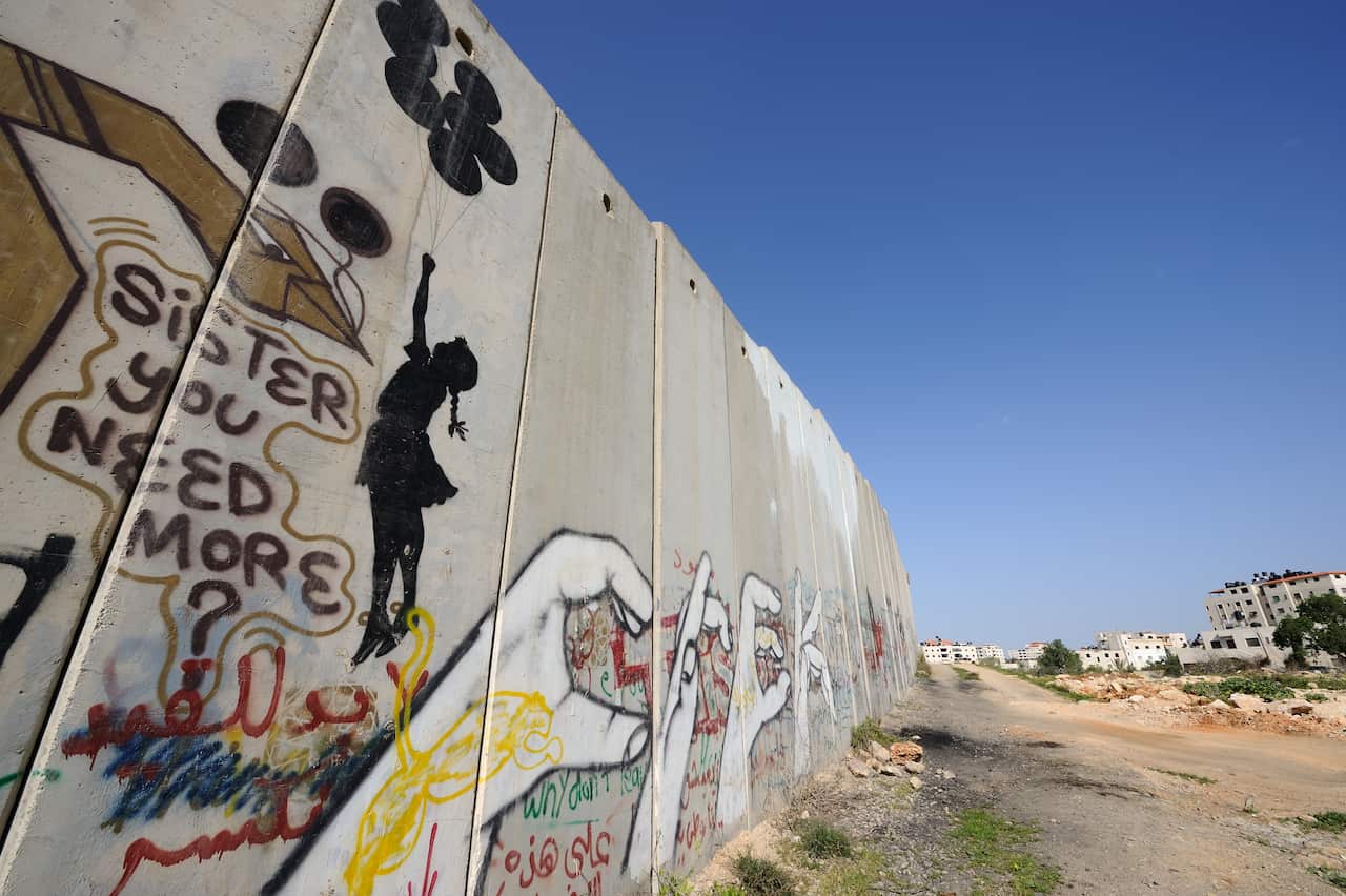 A long concrete barrier. There is artwork on it including four white hands in a row making shapes, and a silhouette of a girl holding up balloons which are lifting her up.