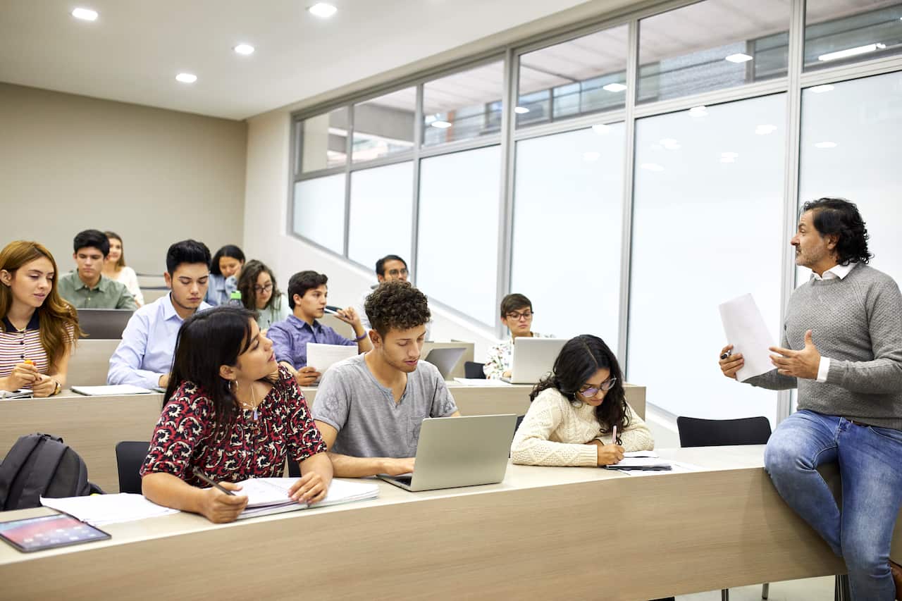 A man dressed in jeans and a grey sweater speaks to students sitting at long bench tables