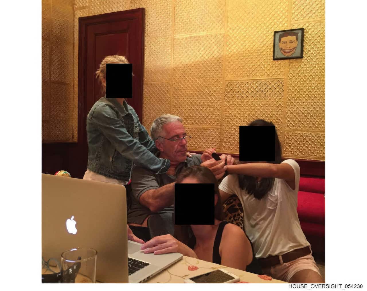 Jeffrey Epstein sits at a table with three young women, whose faces are obscured by black squares, in a room with patterned gold walls and a framed drawing of a smiling face.