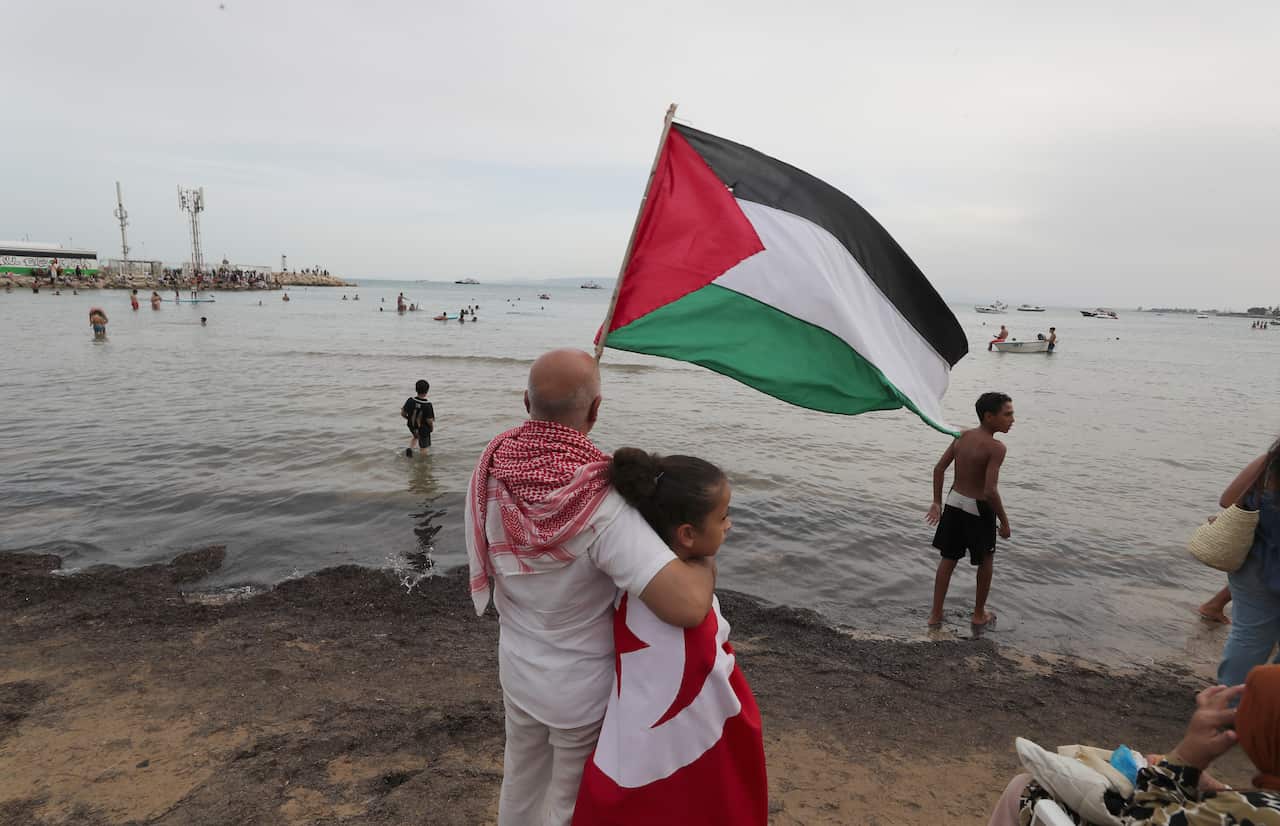 A man waving a Palestinian flag while holding a young girl, standing on a beach.