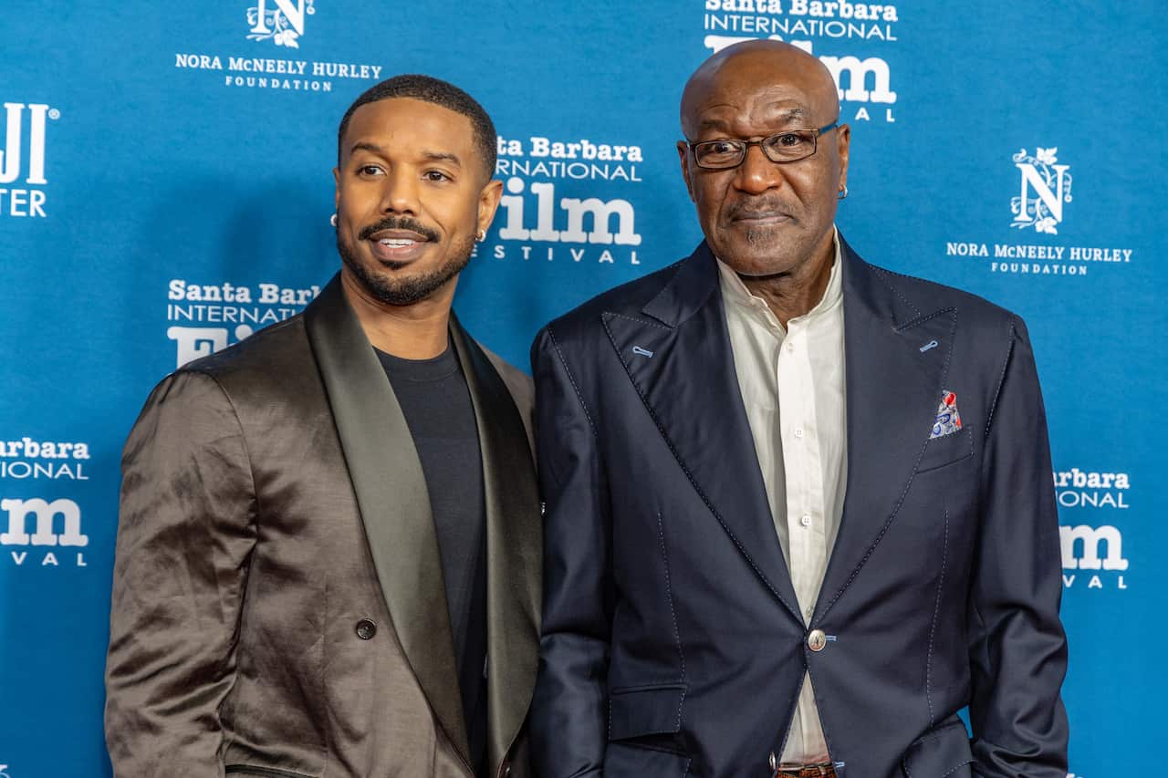 Two men in suits stand in front of a media wall. 