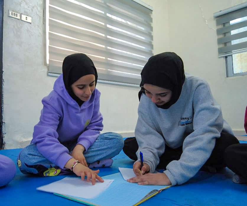Two young girls sit on the ground wearing hijabs and writing in notebook