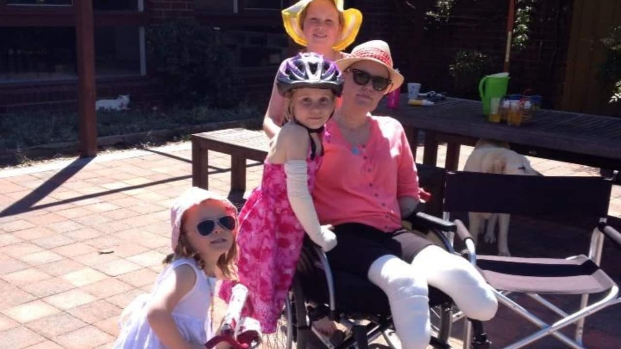 three young girls in pink pose with their mother who is in a wheelchair and has her amputated legs wrapped in bandages. They are in a backyard with a table and a golden Labrador