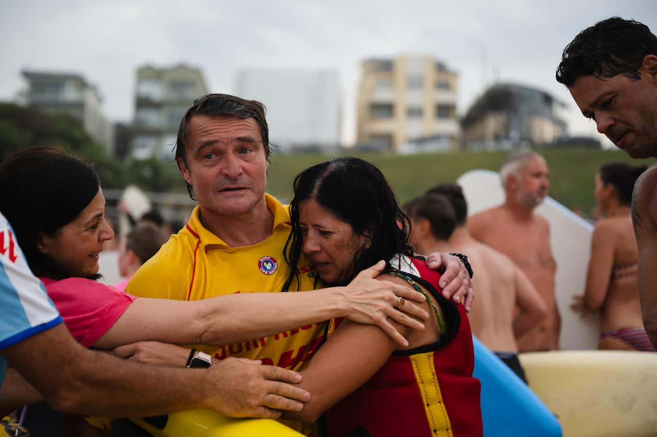 A man and woman embracing each other at the beach, another woman comforts them. 