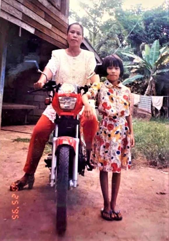 A young girl wearing a floral dress stands next to a woman on a motorbike.