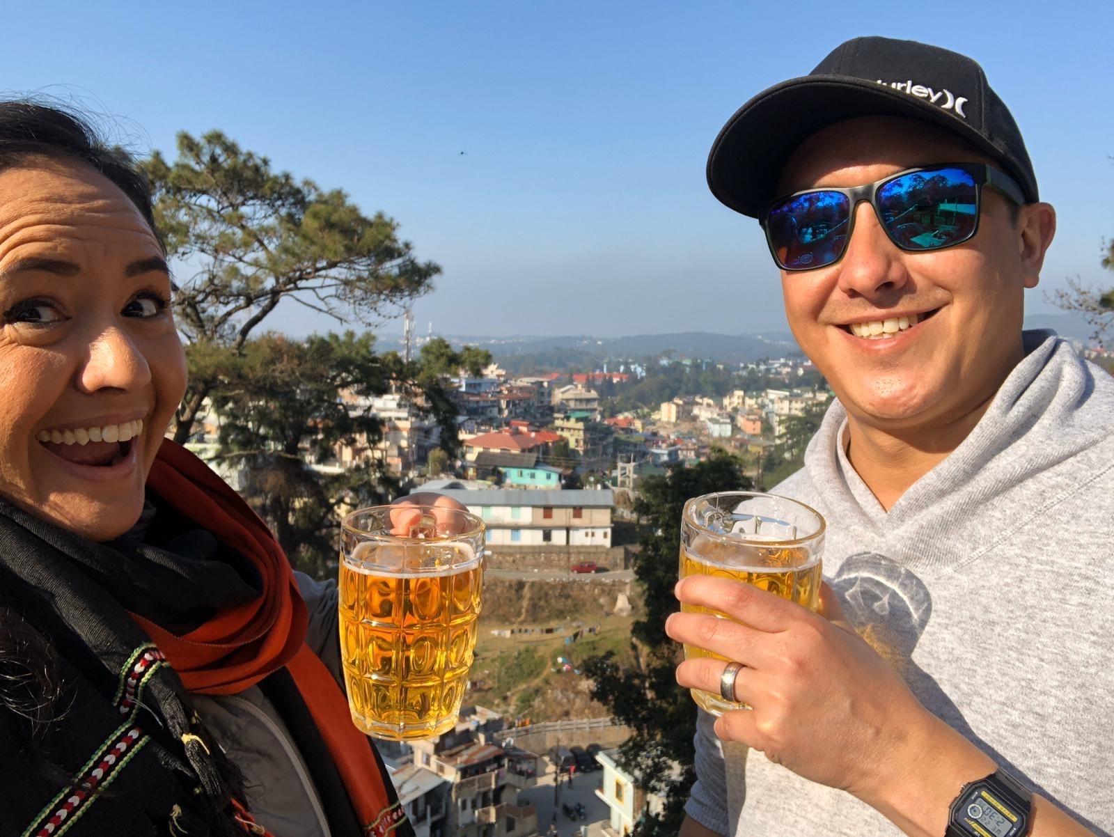 Um homem e uma mulher segurando cervejas e sorrindo, uma cidade ao fundo. 