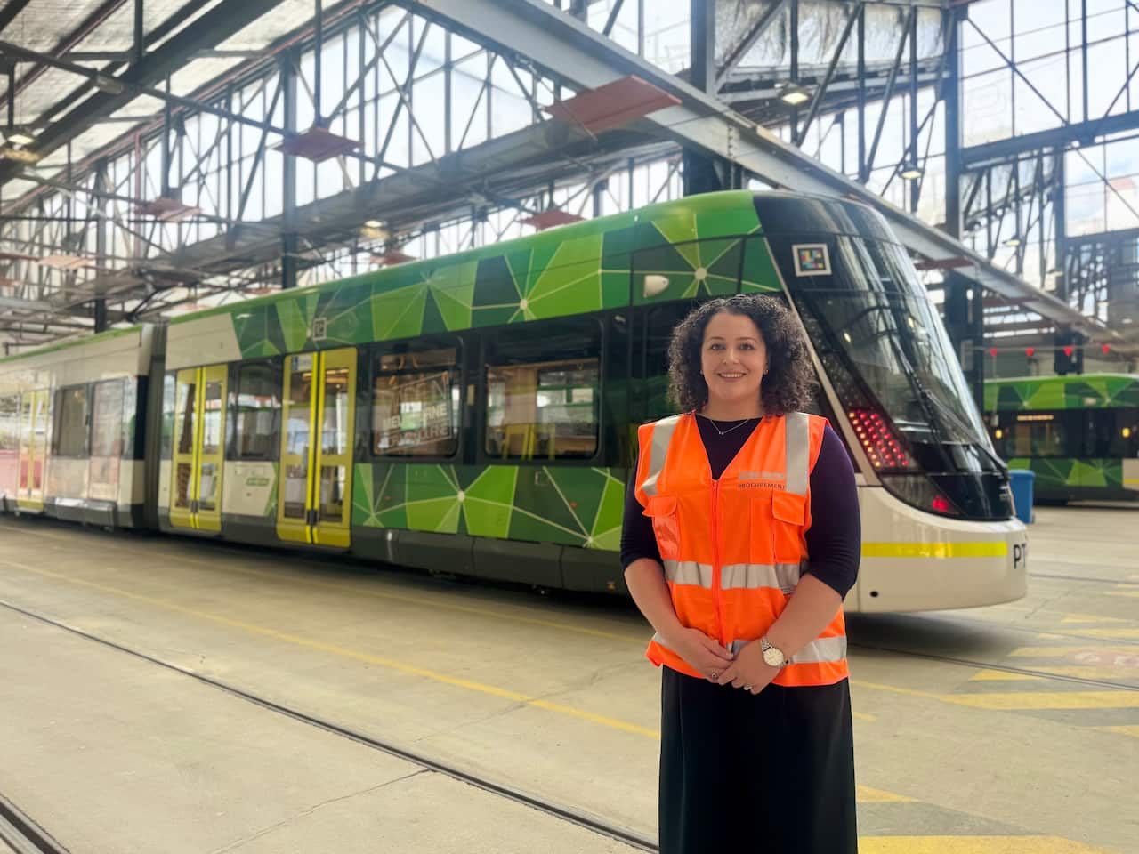 A curly haired woman wearing a hi vis jacket is standing in front of a tram at the depot.