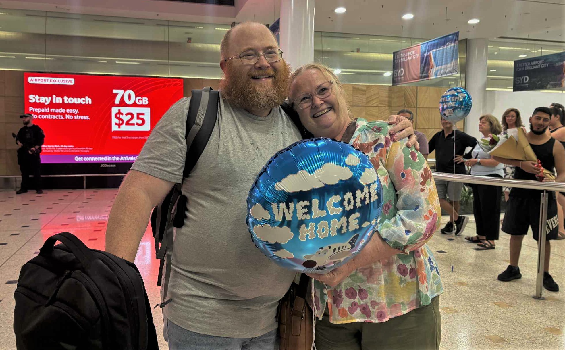 A man in a grey t-shirt with a beard stands on the left embracing a woman in a floral shirt on the right with a blue balloon that reads 'Welcome Home' 