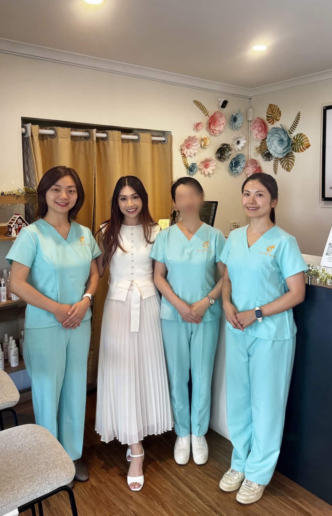 Four women, three in light blue scrubs and one in a white dress, stand together smiling in a well-lit indoor setting decorated with paper flowers.