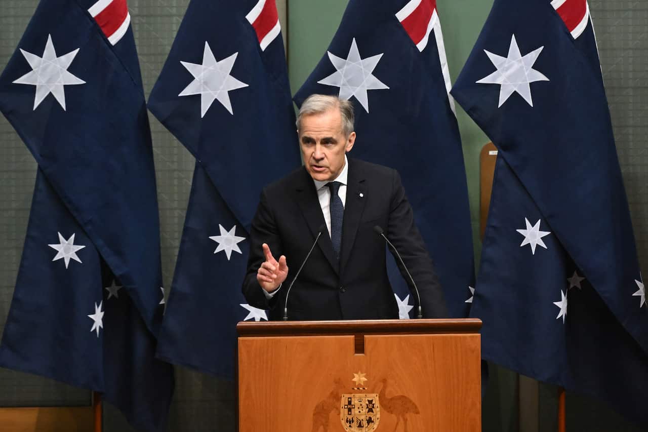 Mark Carney, satnding at a podium and speaking into a microphone, in front of several Australian flags.