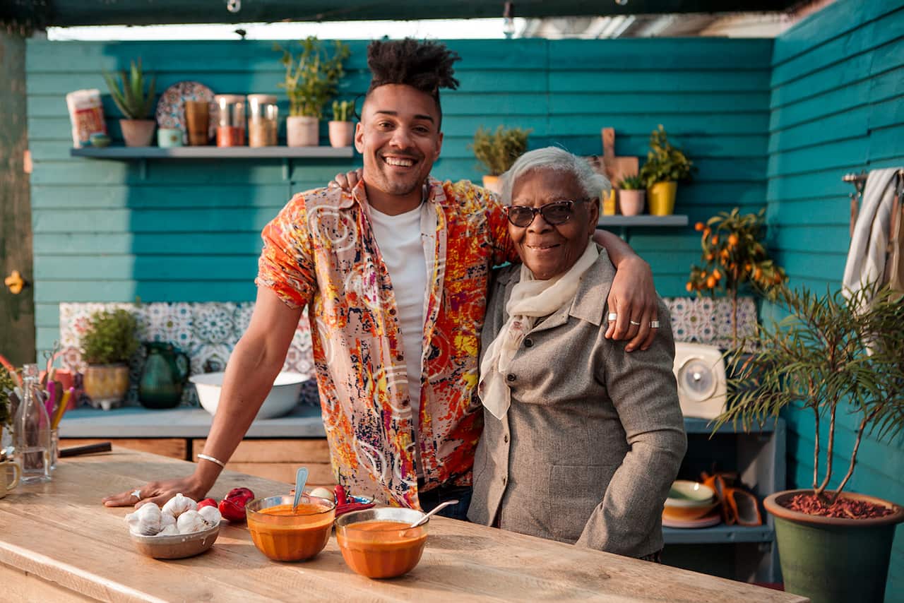 Nico, a tall man with a bright shirt stands in a kitchen, with his arm around his grandmother.