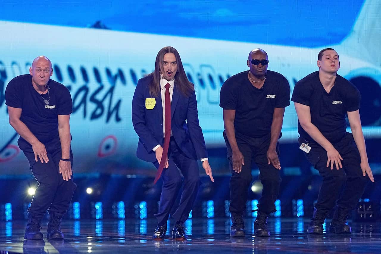 Four men dancing on stage. One has long hair and is wearing a suit
