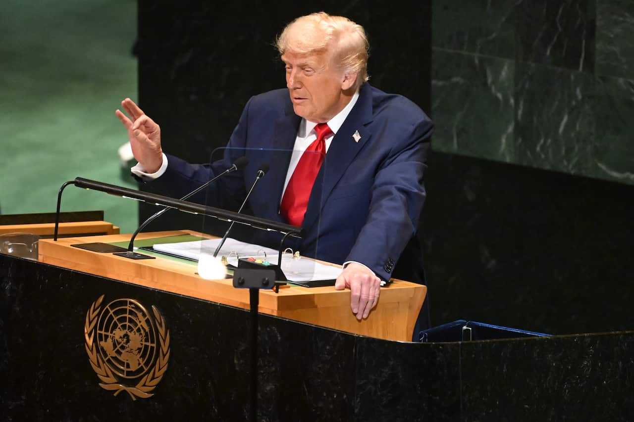 Donald Trump, wearing a dark blue suit and red tie, holds his right hand up with his thumb and forefinger pressed together as he speaks from behind a lectern