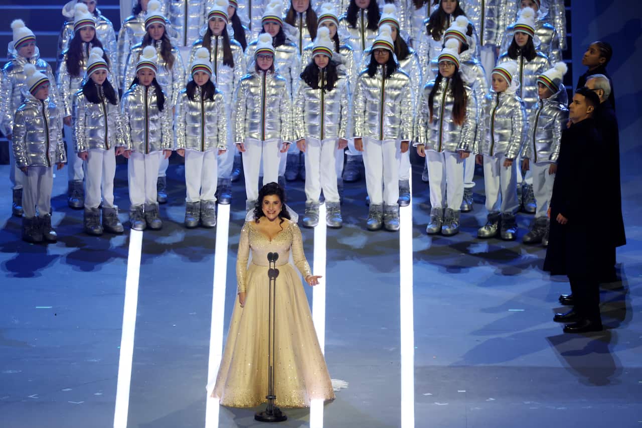 A female singer dressed in a white gown during a performance. Behind her, performers wearing silver puffer jackets and white beanies and pants. 