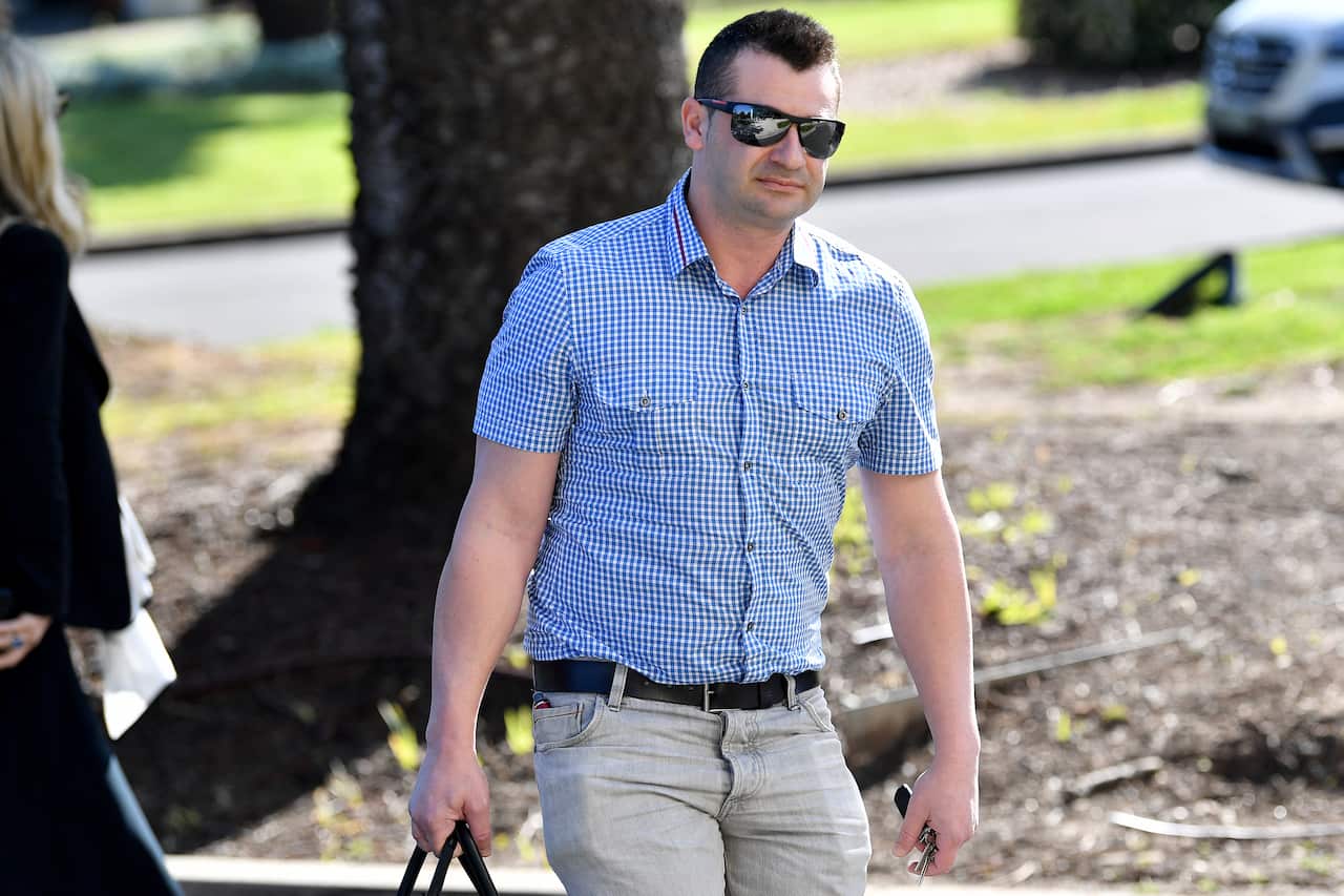 A man wearing a blue check shirt and jeans and carrying a bag walking outside
