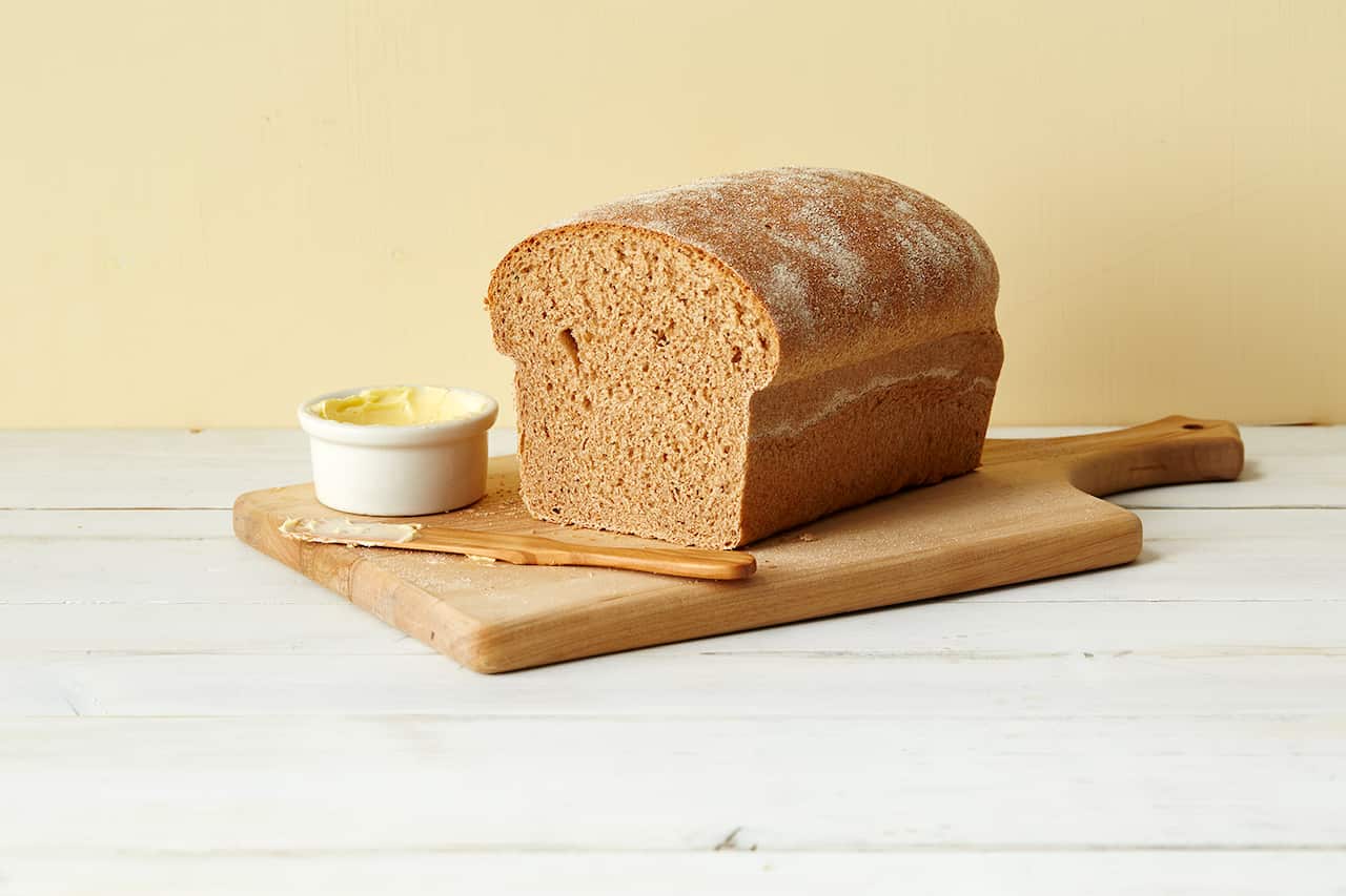 A tall loaf of bread with a wider, rounded top and the front sliced off sits on a wooden board. A pot of butter and a knife sit in front of the bread, on the board.