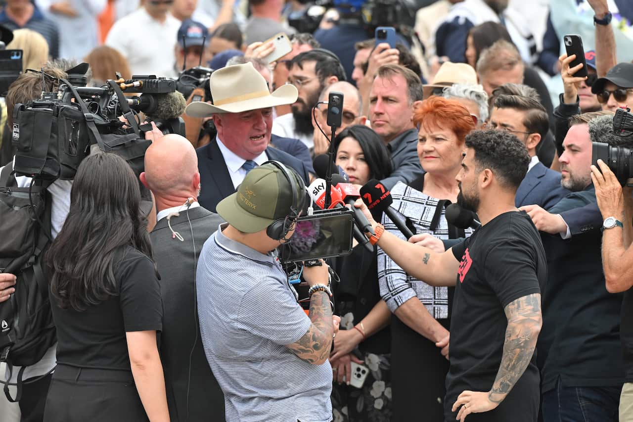 Big crowd of people holding cameras and microphones encircle a man and woman.