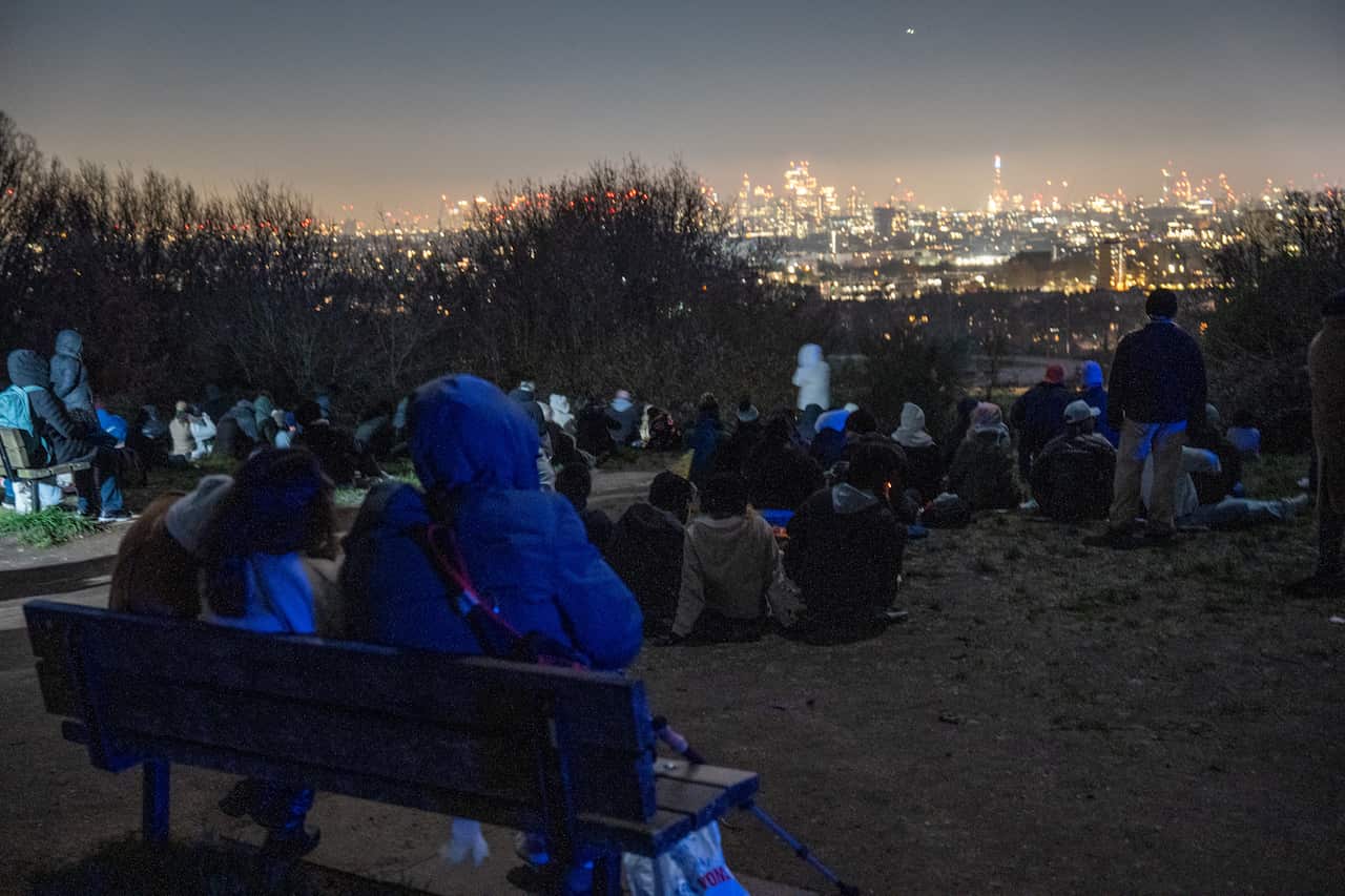 People sitting in a park at night watching a cityscape.