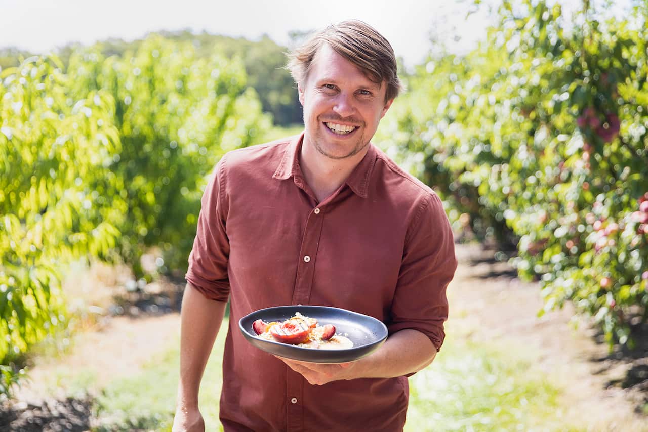 A smiling man stands in an archard, holding a plate with what looks like a stonefruit dish. 