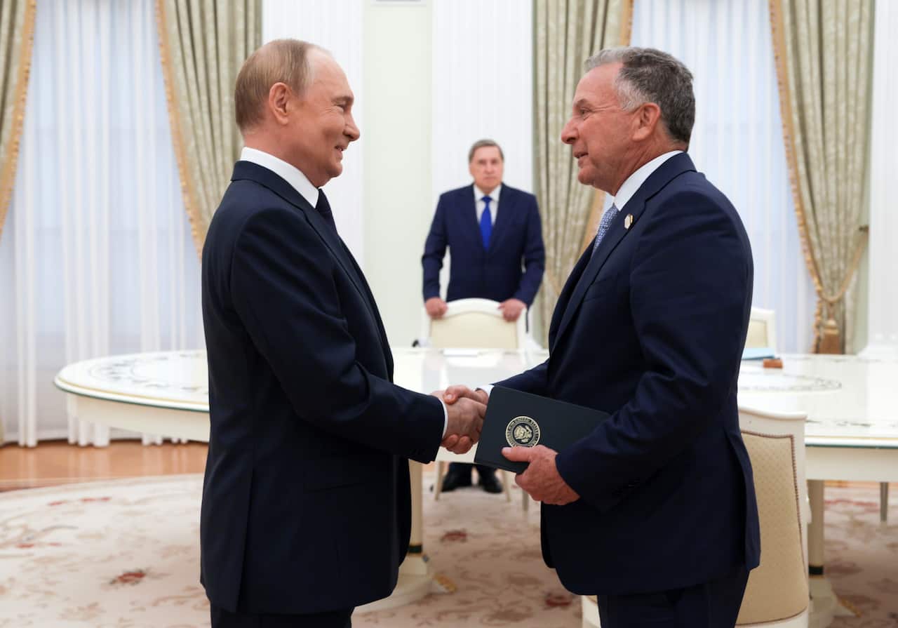Two men in suits shake hands inside a room with another man standing at a distance behind a chair.