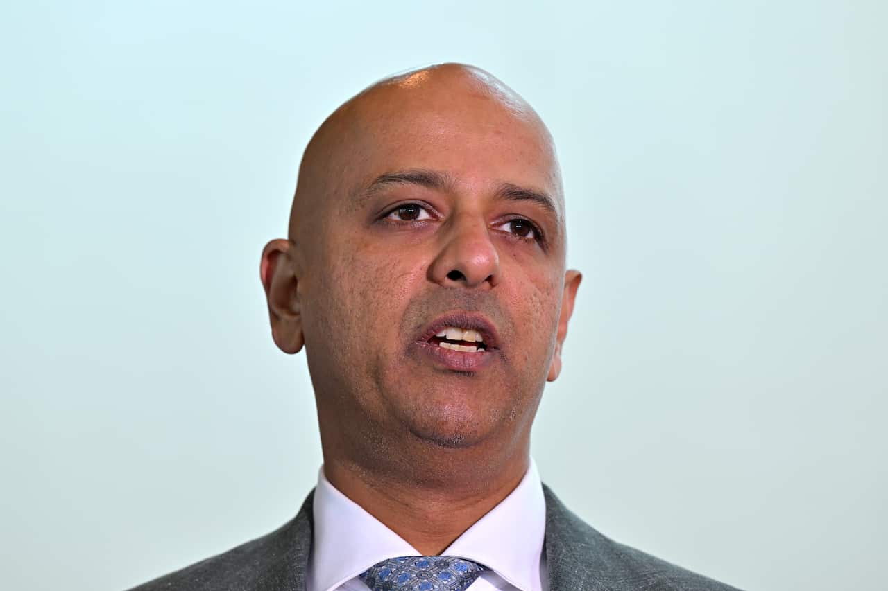 A manwearing a black jacket, white shirt and blue tie speaking against a wall.