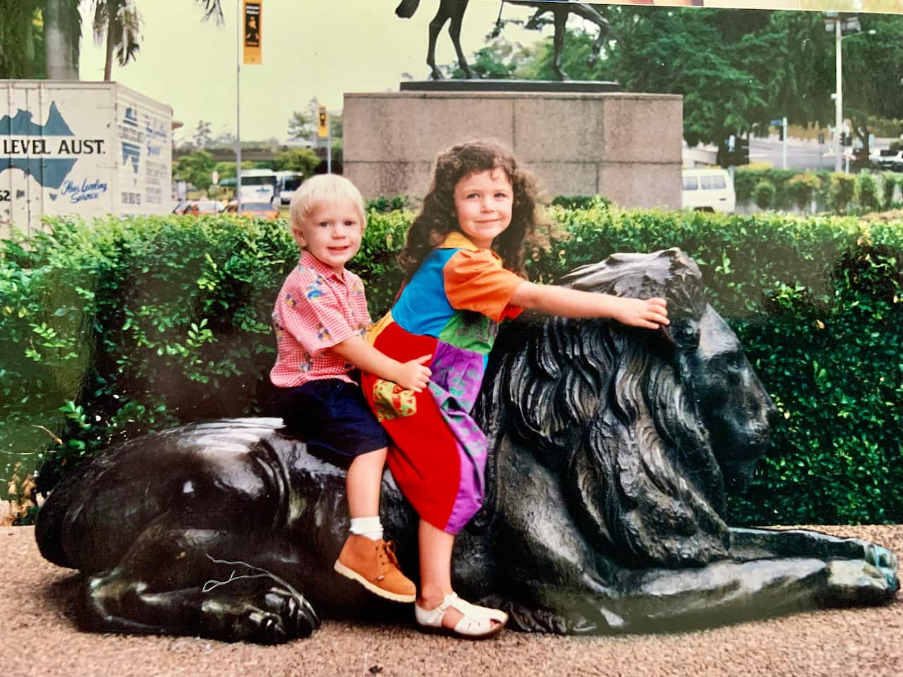 a young boy and girl sit on ground-level a statue of a black lion outdoors