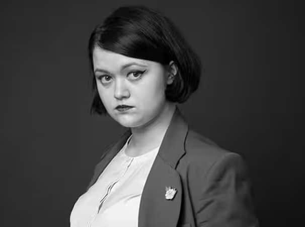 A black and white portrait photo shows a young woman with dark, bobbed hair wearing a blazer, white shirt, and dark lipstick, looking directly at the camera with a serious expression.