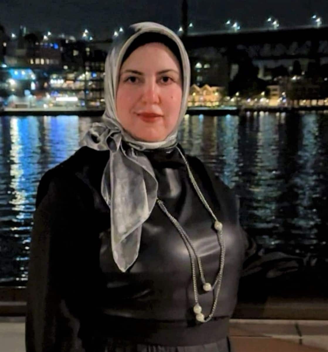 A woman wearing a black dress and a headscarf stands in front of water at night.