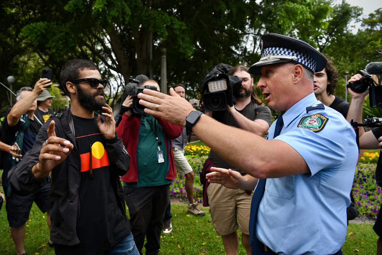 Un oficial de policía australiano señala a un hombre que lleva una camiseta con la bandera aborigen en un parque mientras varios miembros de los medios graban la interacción.
