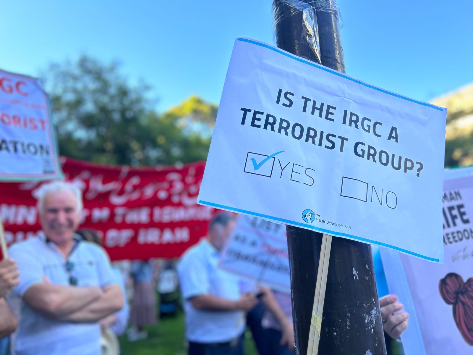 Blurry image of people protesting with banners on a lawn. A poster on a stick in the foreground reads: Is the IRGC at terrorist group? It has yes and no boxes below the question and there is a tick in the yes box.