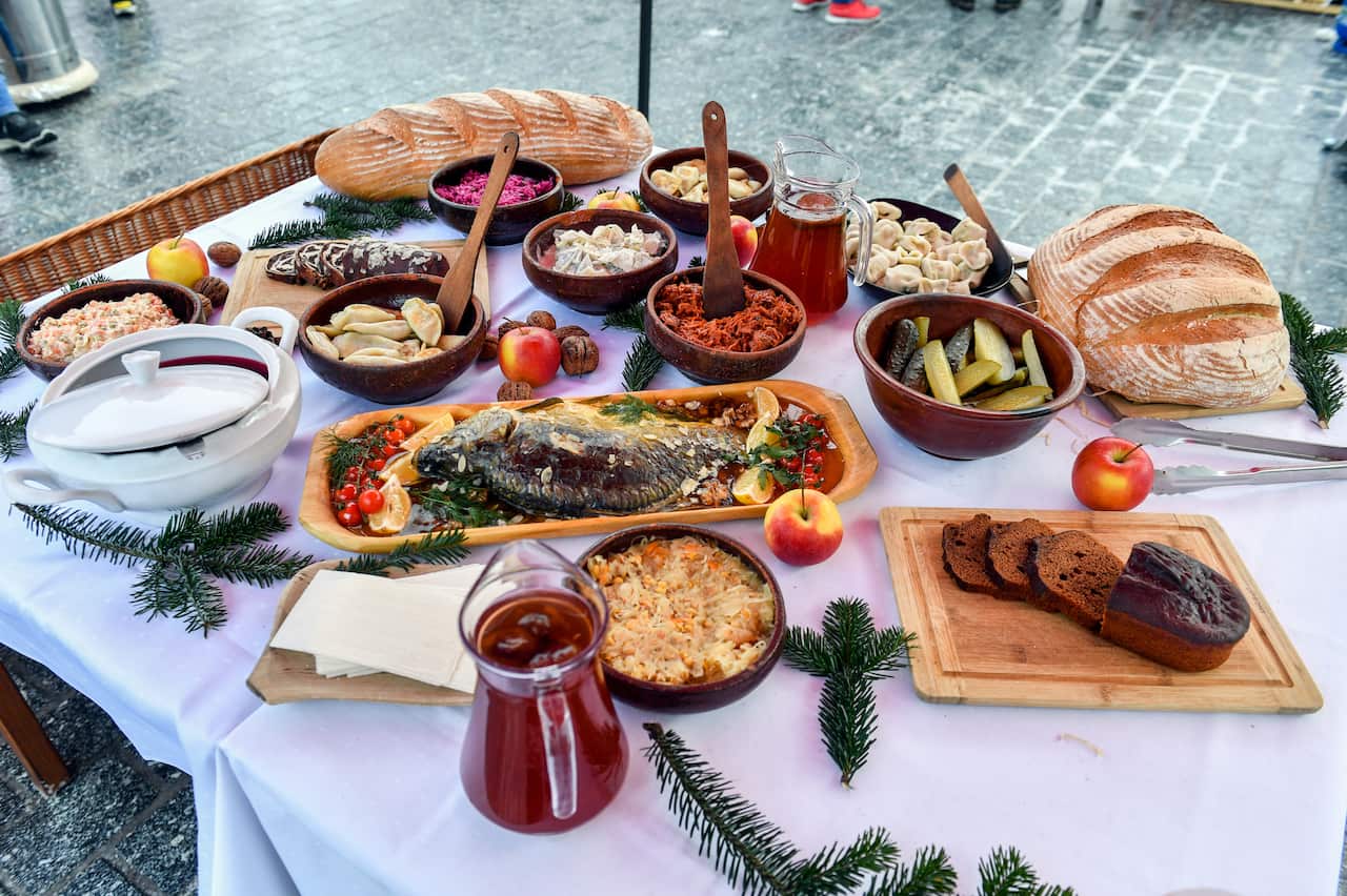 Una mesa al aire libre está repleta de una variedad de platos navideños tradicionales de Europa del Este, que incluyen pescado frito entero, tazones de albóndigas y ensaladas, pan recién hecho y jarras de jugo oscuro.