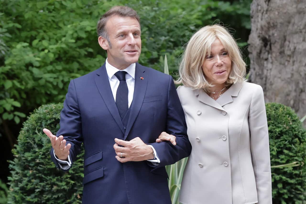 Emmanuel Macron and Brigitte Macron, linking arms, in front of trees and bushes.