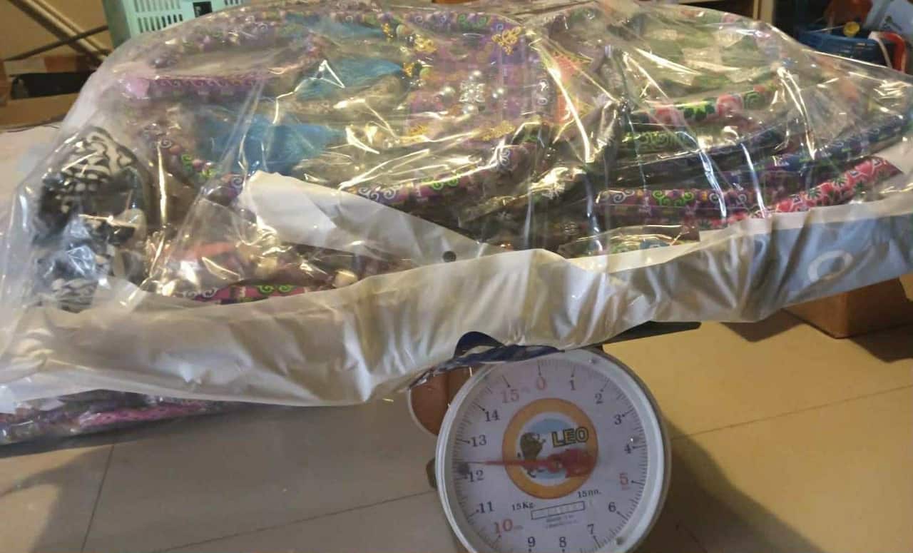 A large, plastic-wrapped package of brightly colored fabric items is being weighed on a round, spring-dial scale that indicates a weight over 15 kilograms.