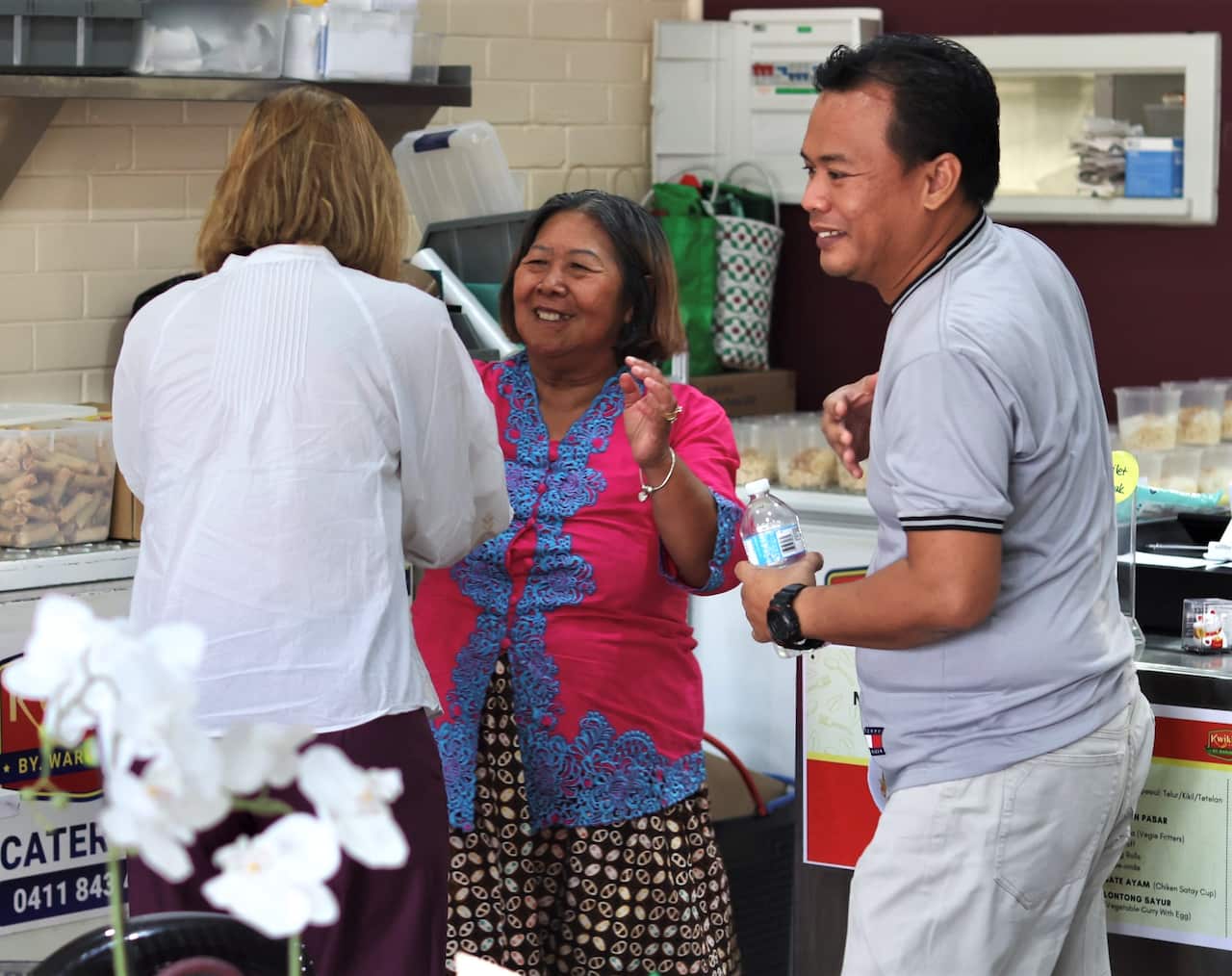 A woman in a pink jacket stands in a cafe kitchen with a man and another woman.