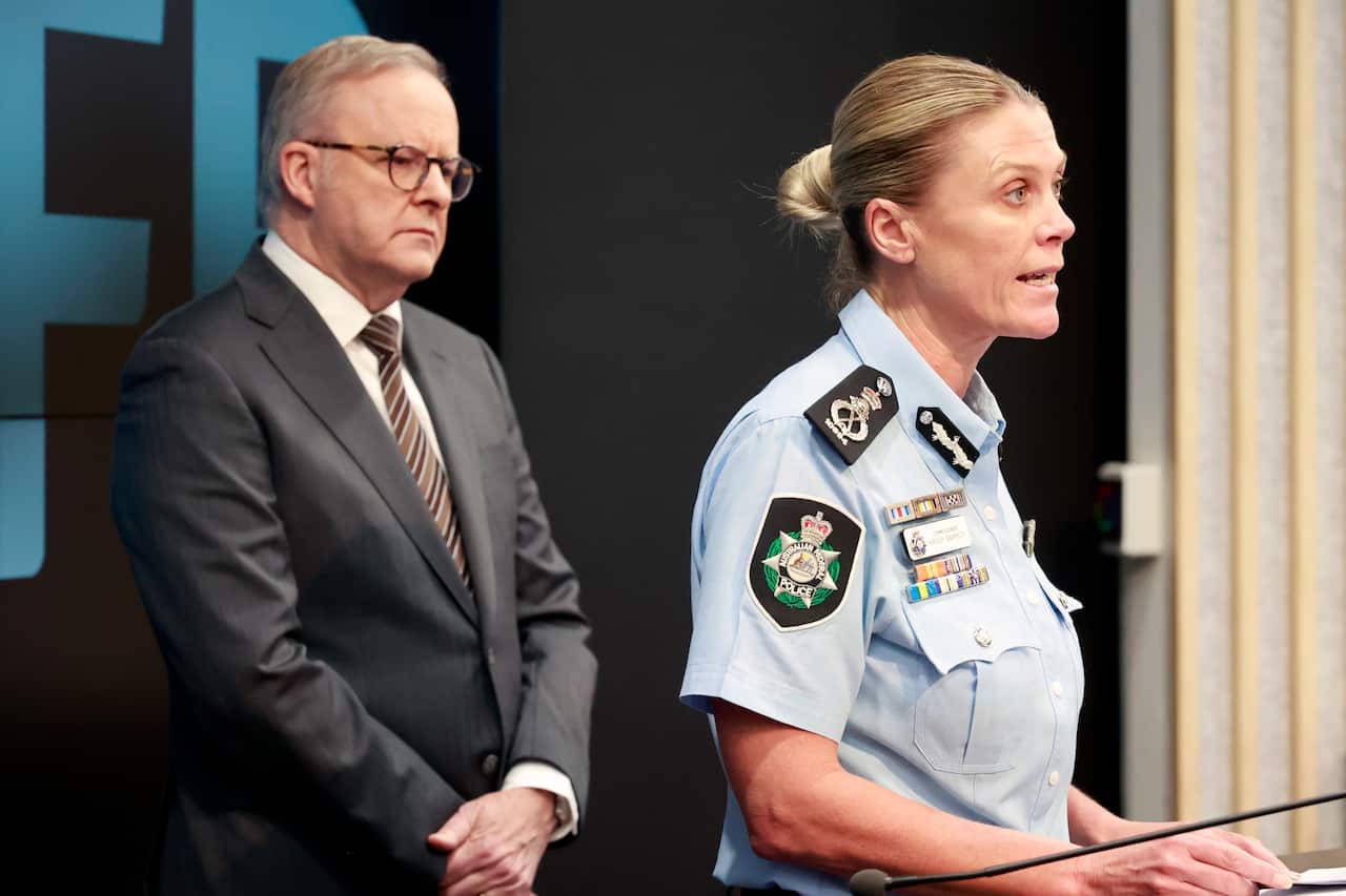 Anthony Albanese, in a black suit, stands next to a woman in a blue police uniform who is speaking at a lectern.