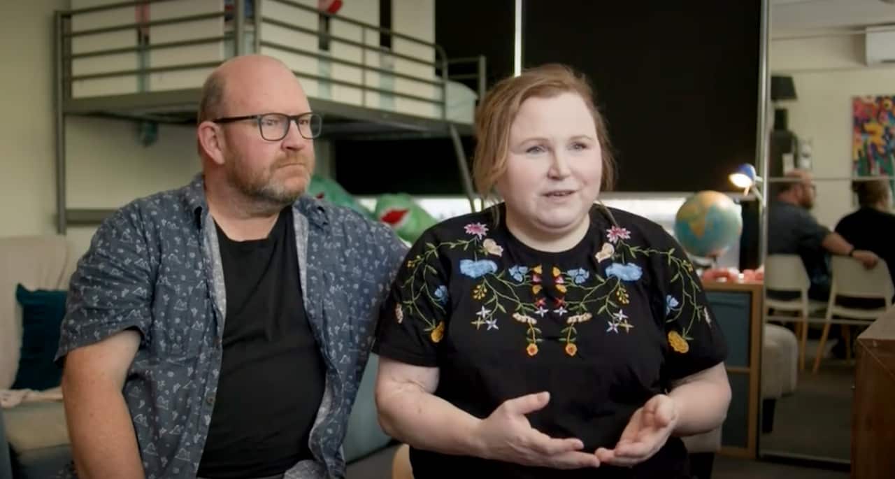 A couple sit inside a cramped room and talk to the camera with a neutral expression on their faces.
