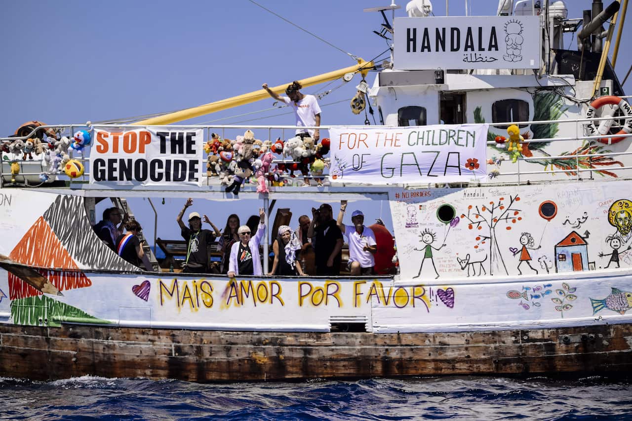 A boat with colourful drawings and signs sets sail with people waving from a distance.