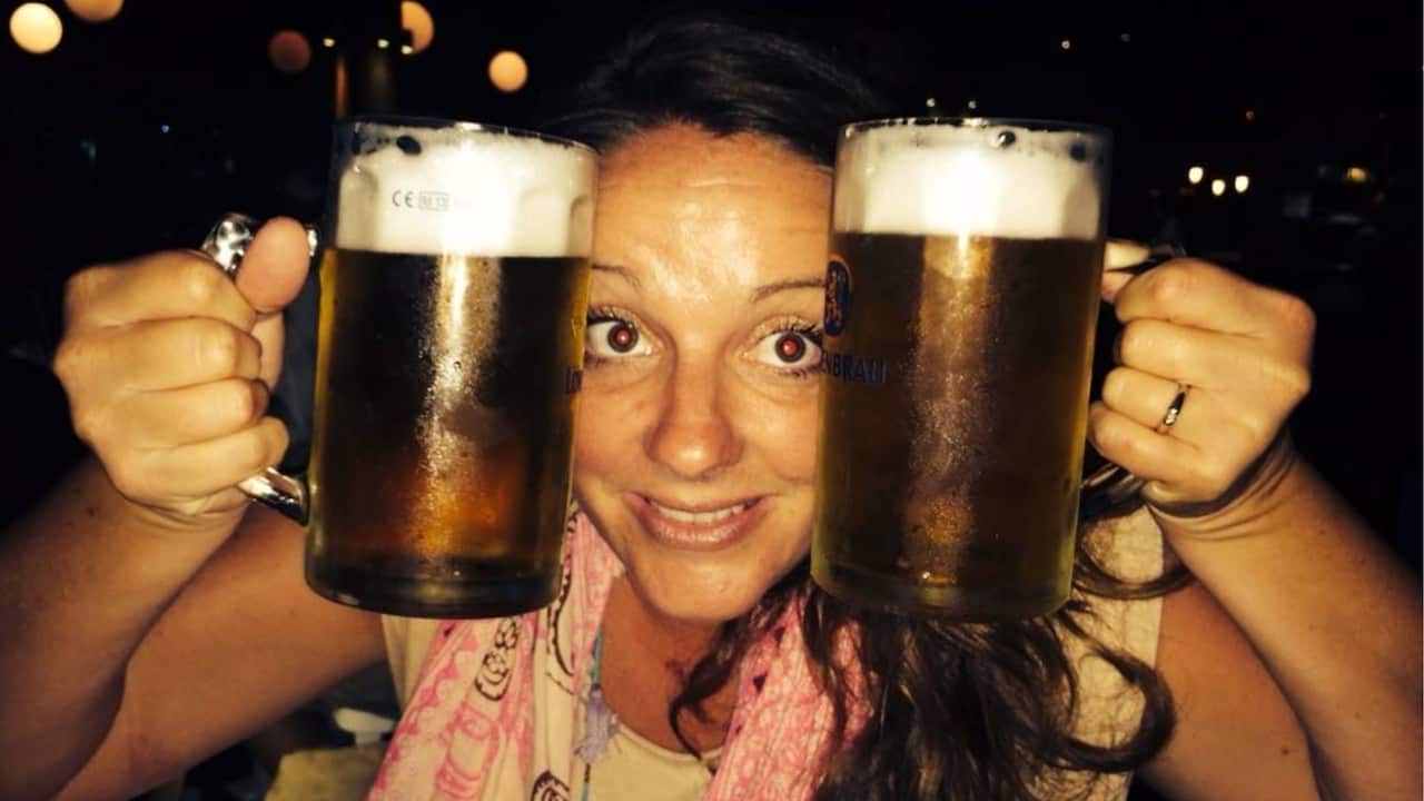 a woman in a pink top on a night out poses holding two pints of beer up either side of her face