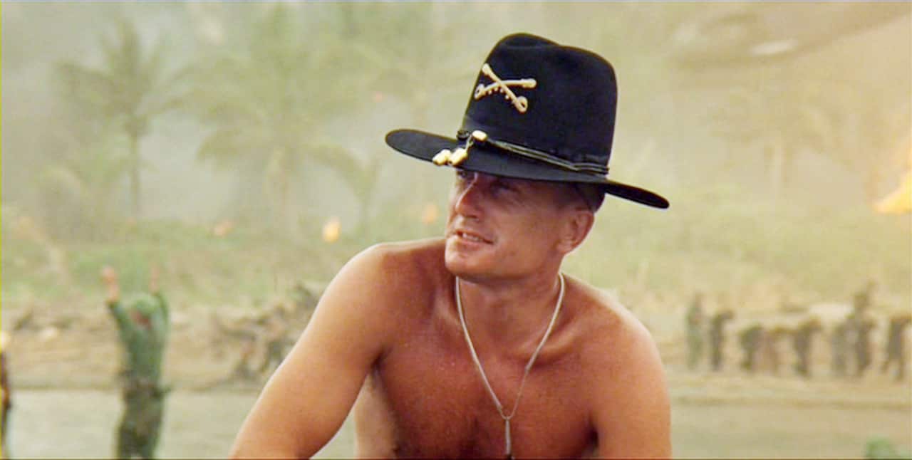 A shirtless man wearing a black cowboy hat, surrounded by soldiers.