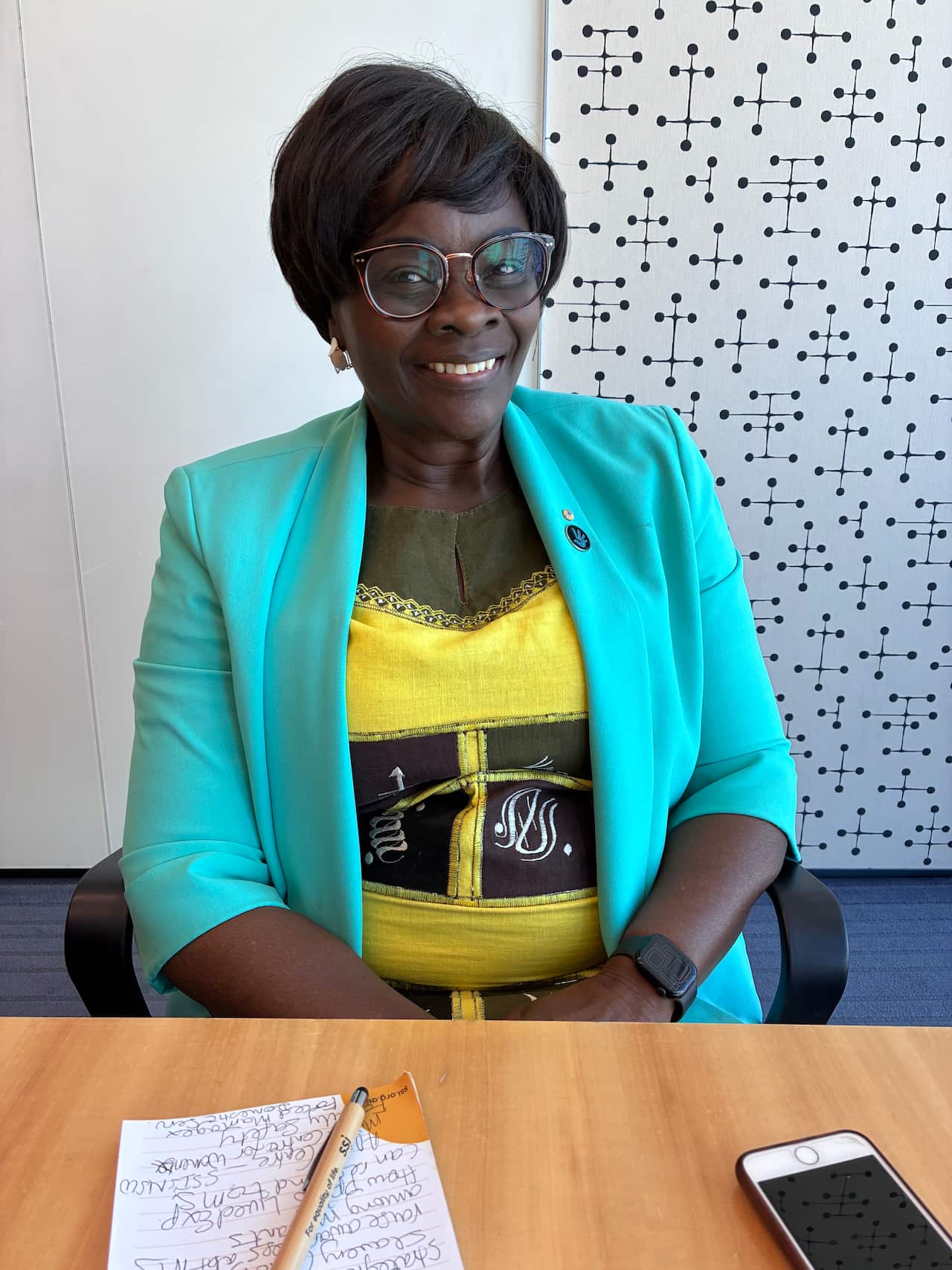 A middle-aged Black woman wearing a yellow dress and turquoise blazer sits at a desk in an office.