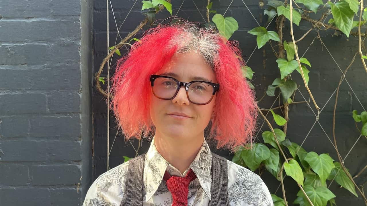 a woman with pink hair and wearing a collared shirt with vest and tie stands in front of a grey mum with vines on it