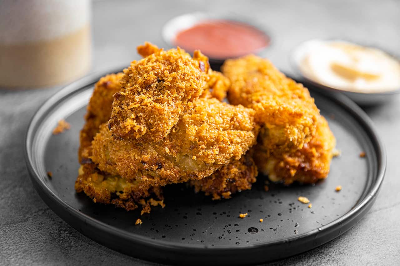 Unevenly shaped golden breadcrumb-covered croquettes sit on a plate. Small bowls of sauce can be seen behind.
