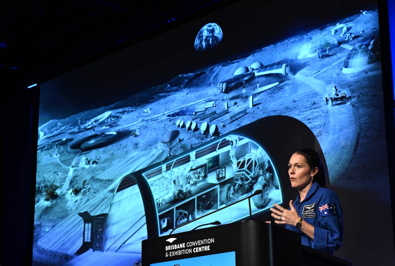 Katherine Bennell-Pegg, wearing an Australian Space Agency flight suit, speaks at a podium at the Brisbane Convention & Exhibition Centre in front of a large screen displaying a concept of a lunar base.