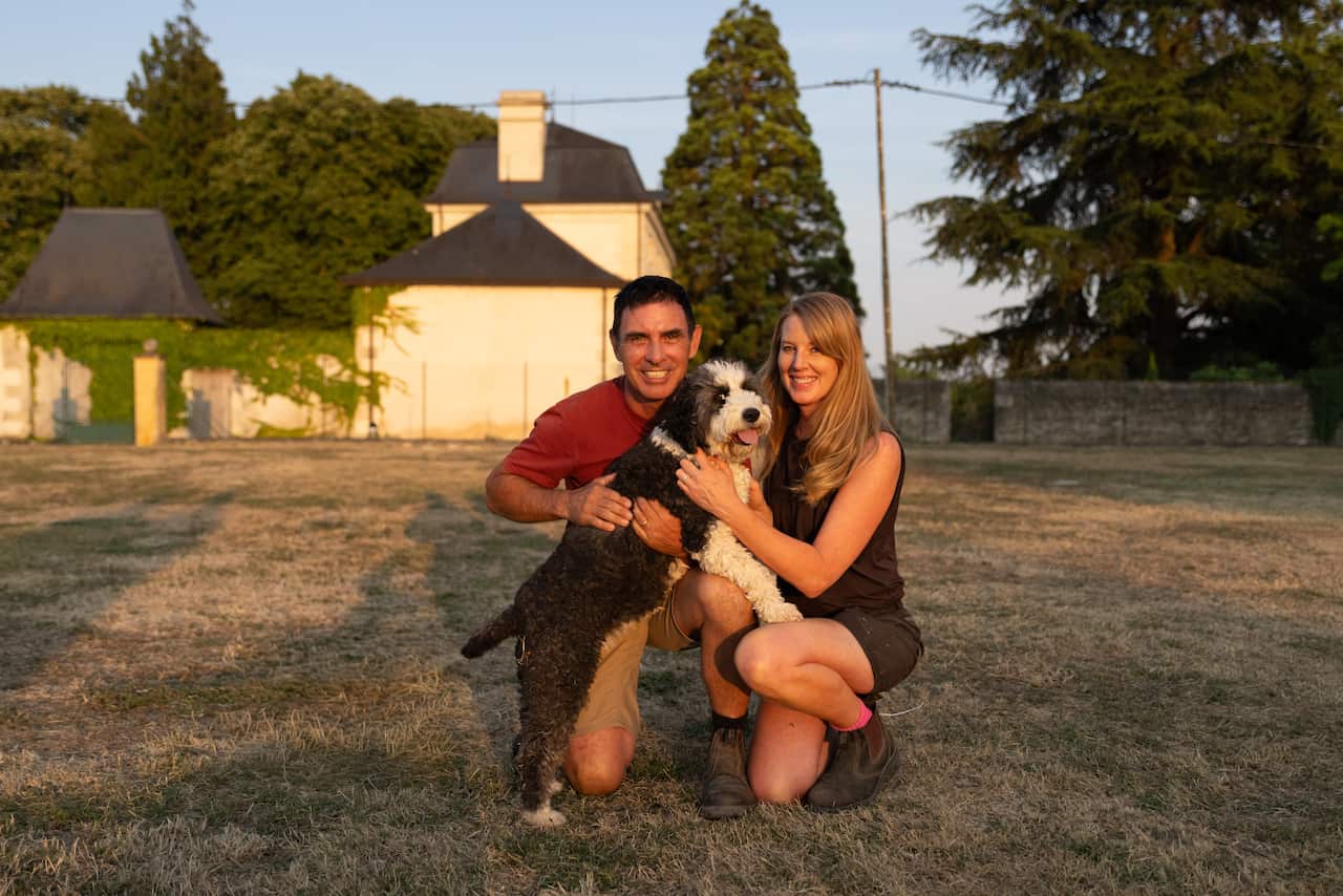 A man and a woman, both smiling, crouch down on a large grassy field. They are holding a large black and white dog. 