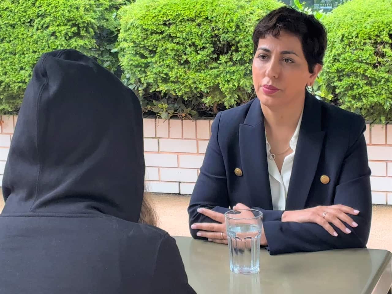 Una mujer con una chaqueta azul está sentada en una mesa con otra persona con una sudadera con capucha negra.